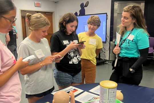 High school students standing around a table using smartphones, talking with a Reliant staff facilitator at a Bite of Reality simulation event.
