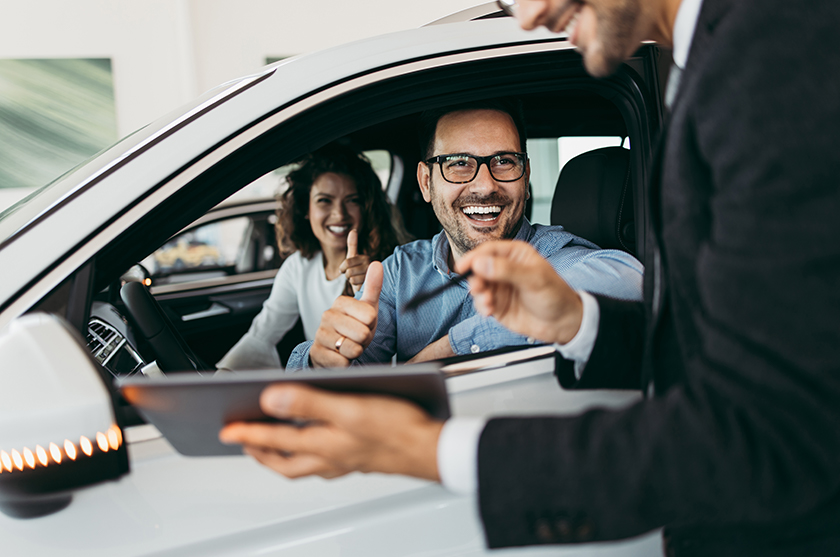 Two people inside car smiling at man standing outside of car holding a tablet device